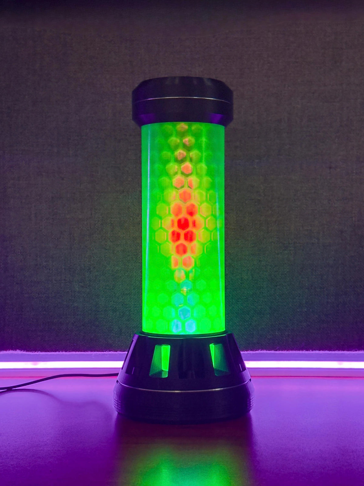 A cylindrical LED light fixture with a black base and top, featuring a glowing green and red honeycomb pattern on a dark textured background.
