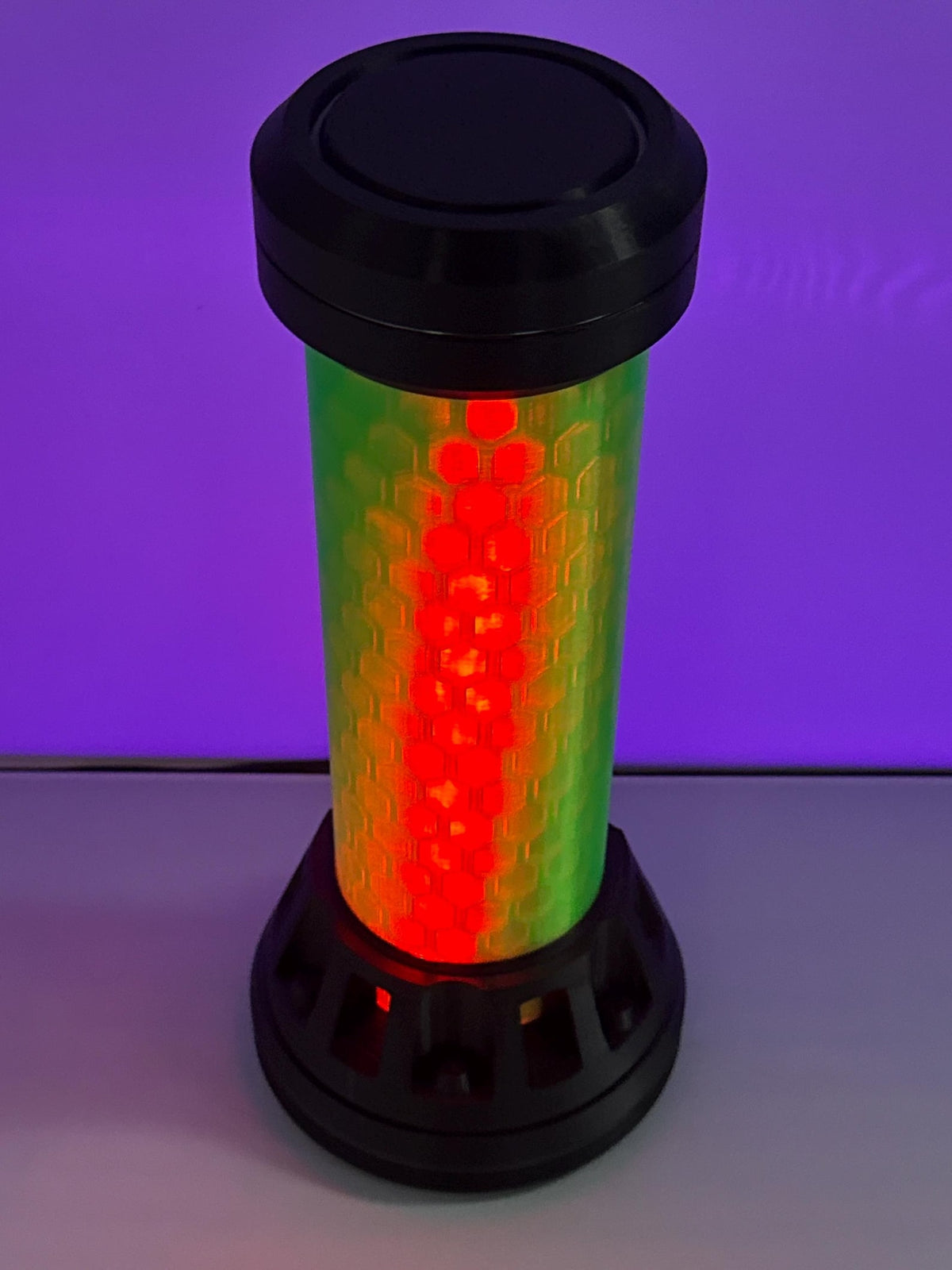 A cylindrical signal light tower with a black plastic top and base, featuring a glowing green-to-red gradient light bar in the center, set against a purple-lit background.