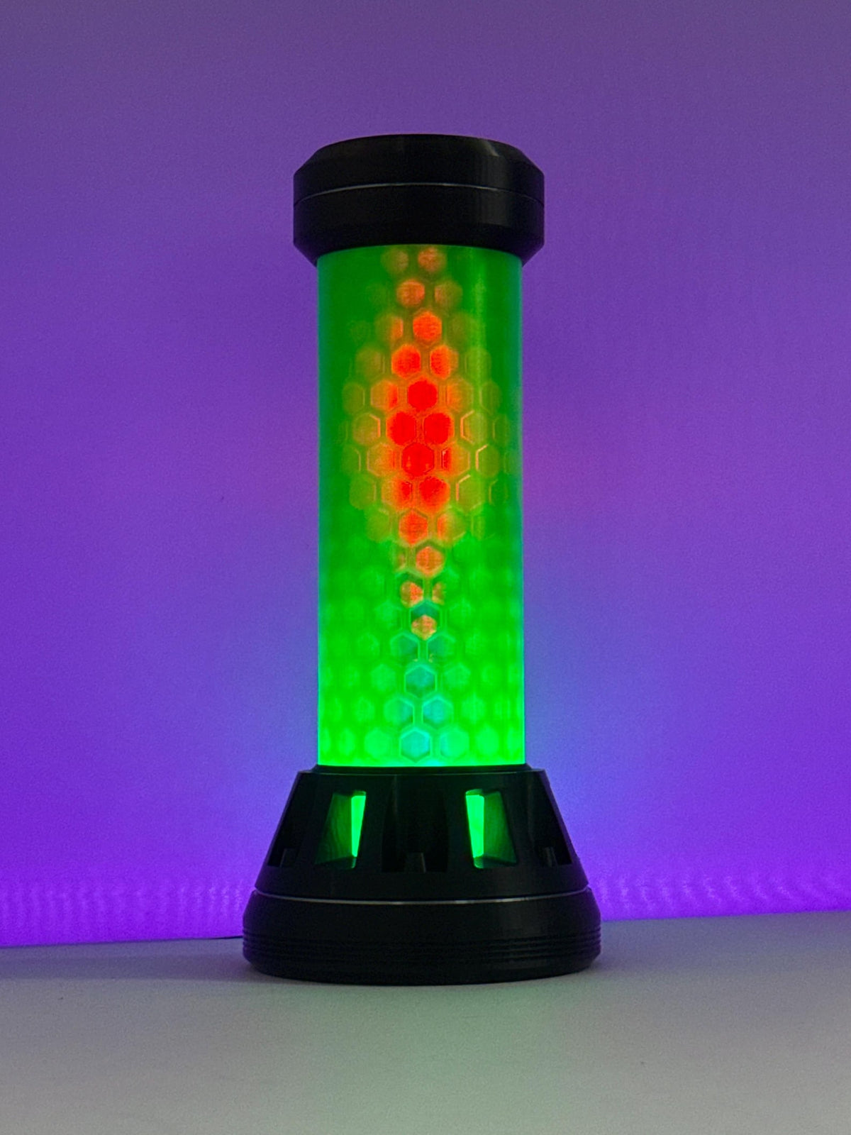 A cylindrical LED light with a black base and top, featuring a green honeycomb-patterned body that glows with a red and orange vertical gradient, set against a purple gradient background.