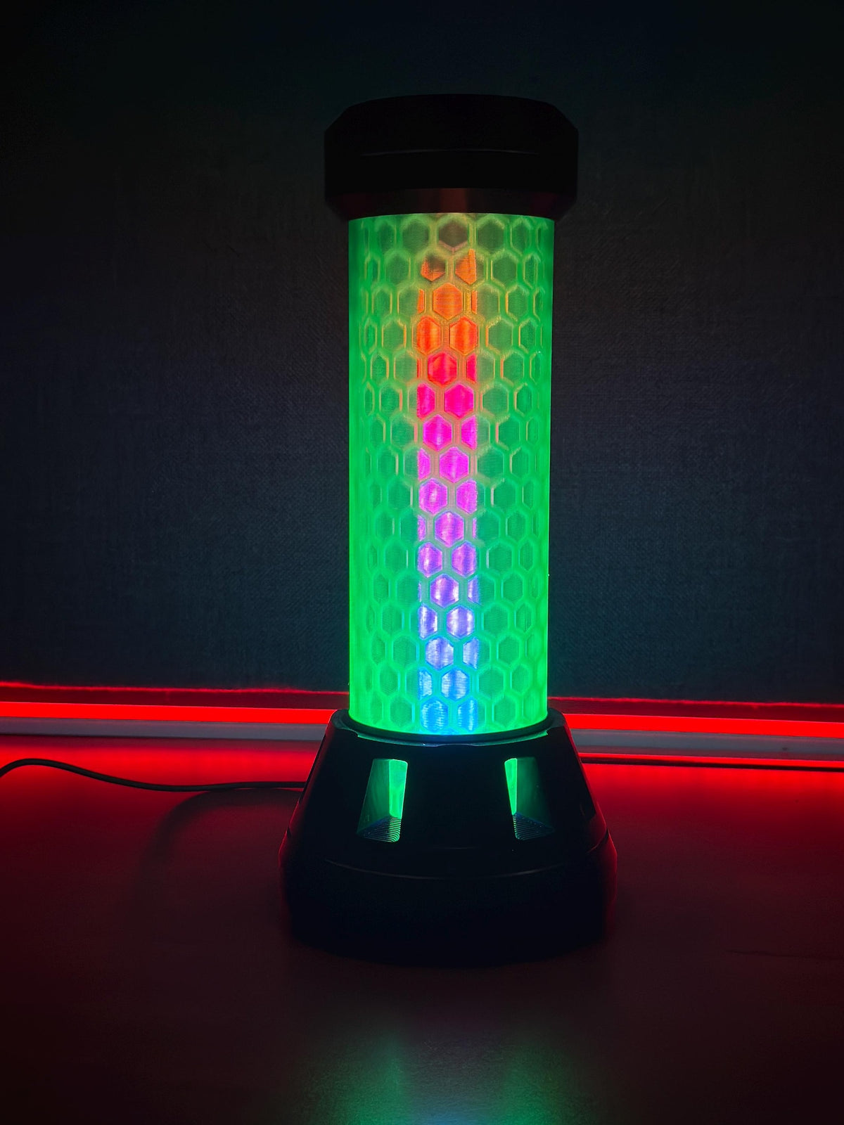 A cylindrical LED light bar with a black base and top, featuring a honeycomb-patterned body that glows with vertical rainbow colors including green, blue, purple, and orange, set against a dark background with red ambient lighting.