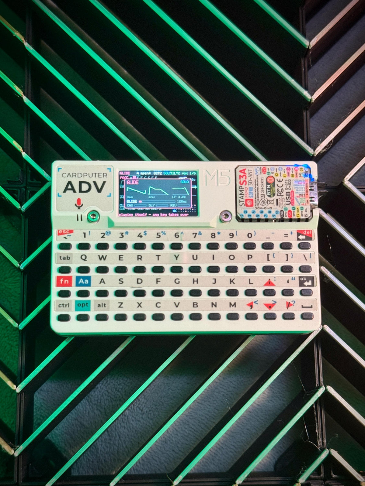 A retro-style electronic cardputer machine labeled 'ADV' with a small screen, printed keyboard, and a colorful circuit board attached on the right, photographed against a black slatted background with green neon lighting.